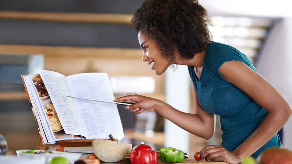 Een vrouw leest een kookboek op een houten standaard in een keuken. Er liggen groenten op een houten snijplank.