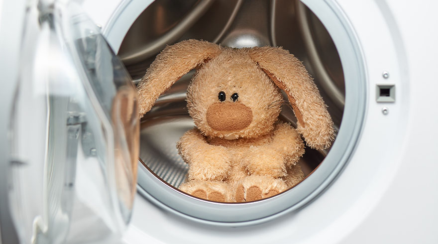 Een knuffelkonijn zit in een witte wasmachine met een open deur, klaar om gewassen te worden.
