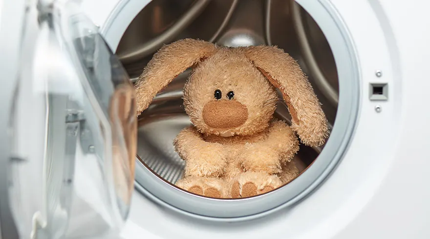Een knuffelkonijn zit in een witte wasmachine met een open deur, klaar om gewassen te worden.