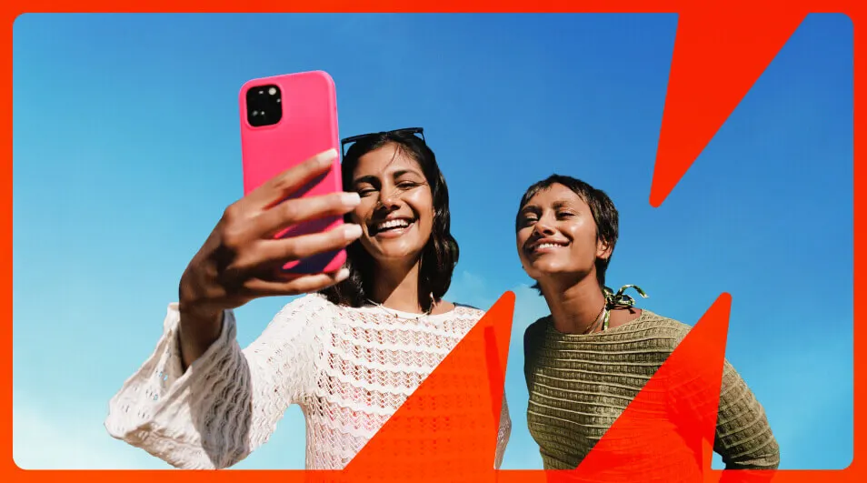 Twee vrouwen lachen en maken een selfie met een roze telefoon tegen een helderblauwe lucht met rode grafische vormen.