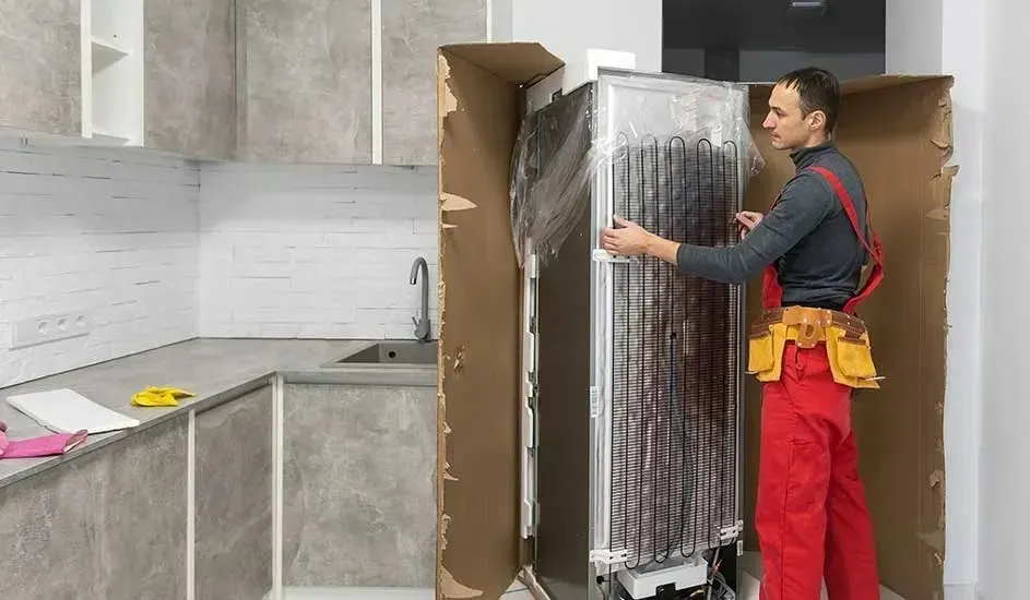 Een monteur in rode werkbroek en gereedschapsgordel plaatst een nieuwe koelkast in een moderne keuken met grijze kasten en witte tegelwand.