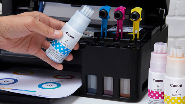 Een hand houdt een blauwe Canon-inktfles boven een printer met blauwe, roze en gele inkthouders. Kleureninktflessen staan rechts.