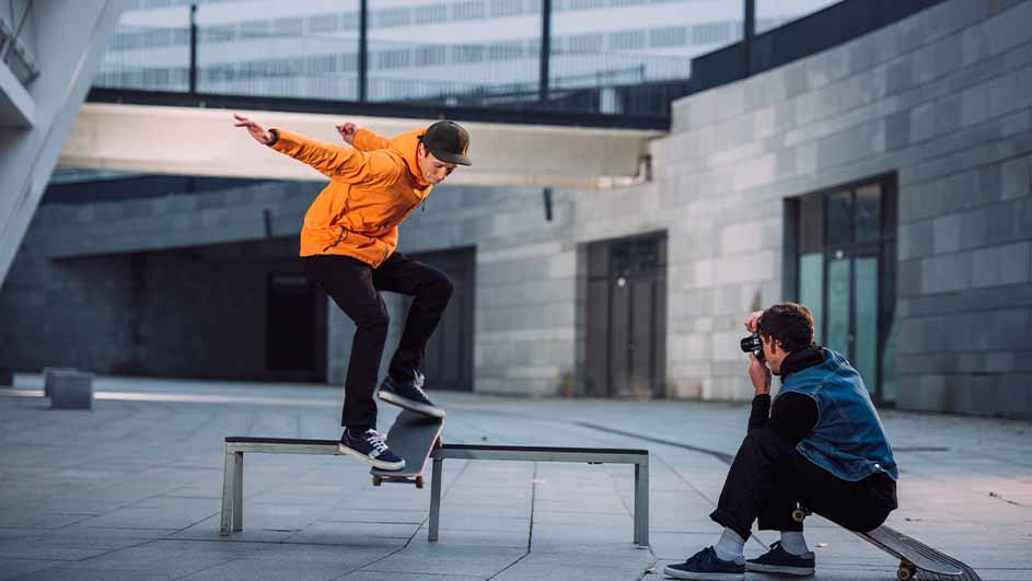 Een persoon in een oranje jas skate over een metalen rail. Een ander persoon maakt een foto met een camera.