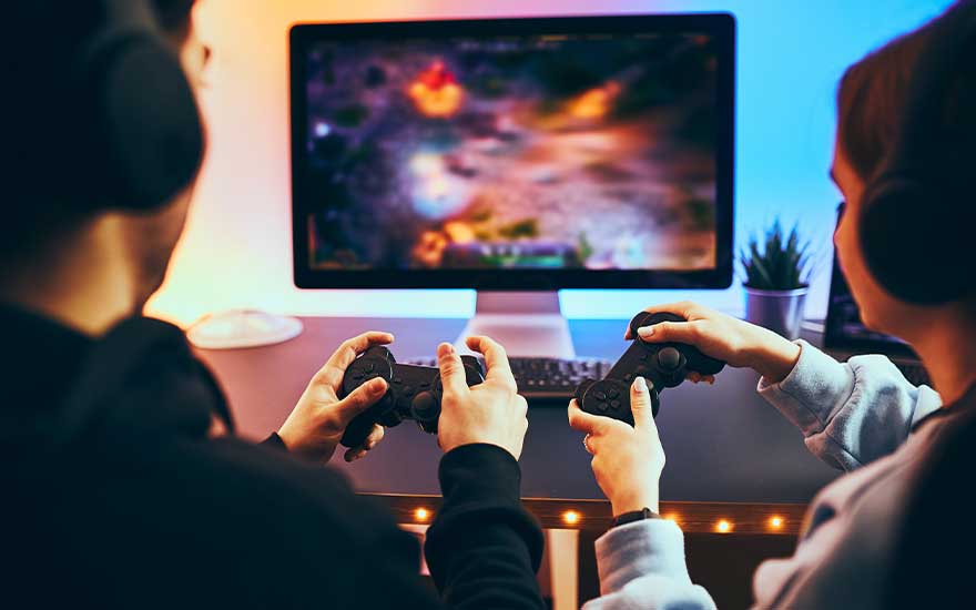 Twee mensen zitten voor een beeldscherm en spelen samen een computerspel, elk met een controller en met hoofdtelefoons op.