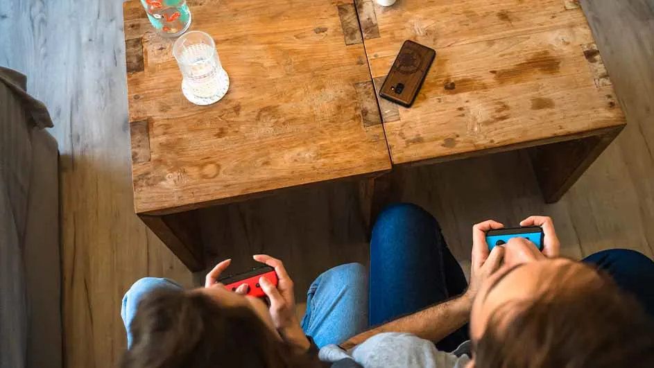 Twee personen spelen videospelletjes met controllers voor een houten tafel met een telefoon en glazen bekers erop.