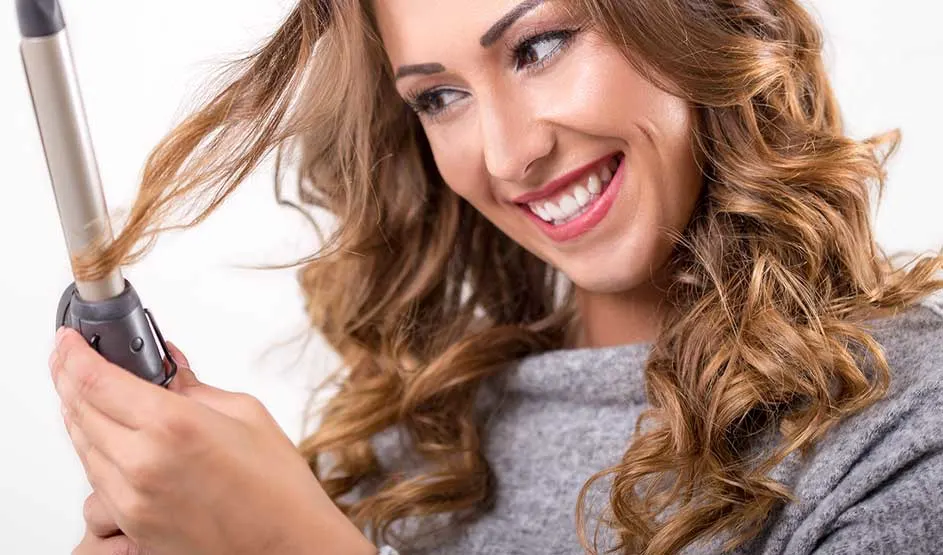 Smiling woman with wavy brown hair using a curling iron to style her locks against a white background.