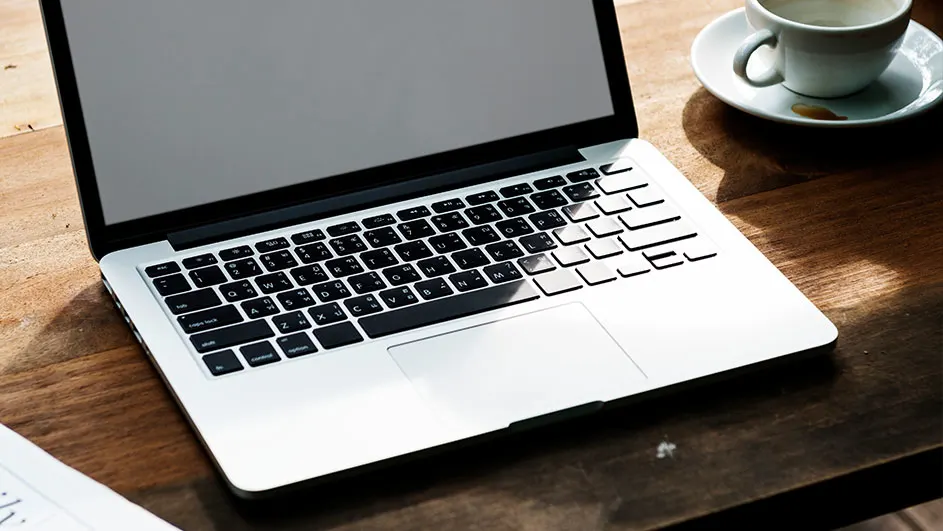 Een laptop met een zilveren behuizing staat op een houten tafel, met rechtsboven een witte kop en schotel.