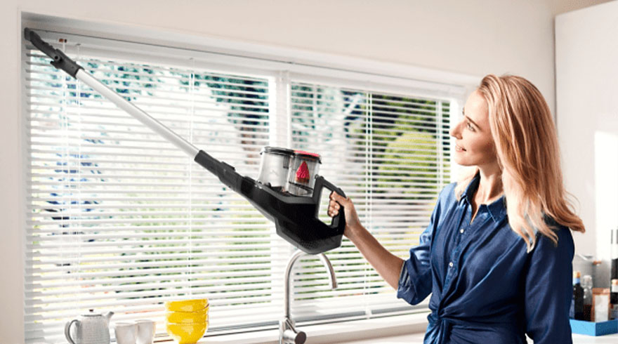 Vrouw stofzuigt witte jaloezieën met een steelstofzuiger. Op de aanrecht staat een theepot en gele kommen.