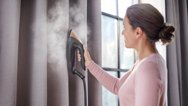 Vrouw stoomt grijze gordijnen met een zwart stoomstrijkijzer. Er komt stoom uit het strijkijzer. Achtergrond: raam.
