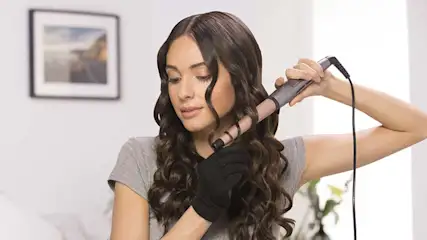 Woman with long dark brown curls styles her hair using a rose gold curling wand, wearing a black heat glove as she neatly wraps a strand around the barrel.