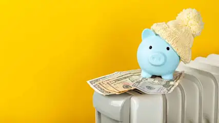 Een blauw spaarvarken met muts op een stapel bankbiljetten bovenop een witte radiator, tegen een gele achtergrond.