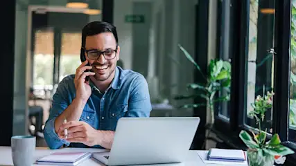 Man zit lachend aan een bureau met een laptop en een telefoon aan zijn oor, in een kantoor setting.