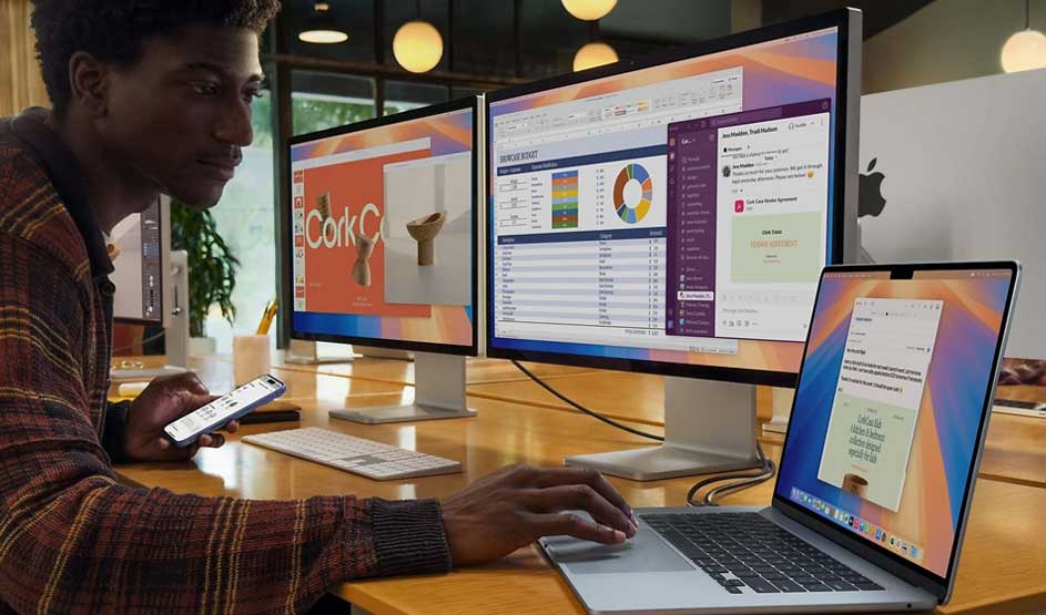 Person working at desk with MacBook and two external monitors displaying data dashboards and charts in modern office setting.