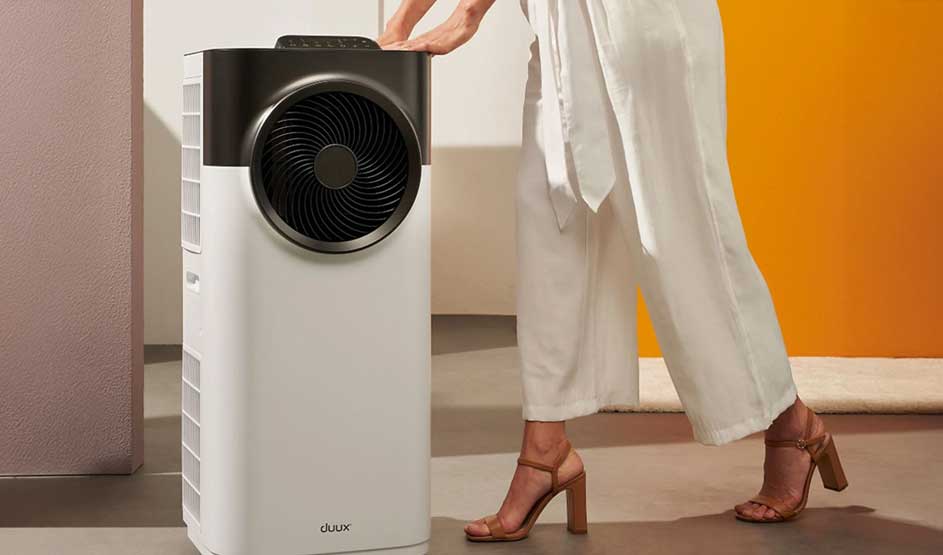 A woman pushes a Duux air conditioner in a modern room with an orange wall and light flooring.
