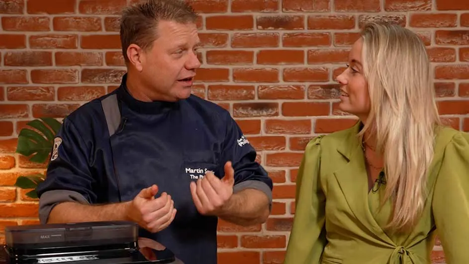 Kok in donkerblauwe koksjas praat met een vrouw in een groene blouse voor een muur van rode bakstenen.