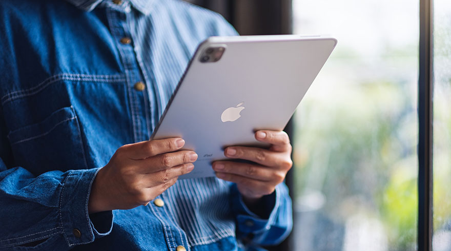 Een persoon houdt een zilverkleurige tablet vast met een appel logo, gekleed in een blauw spijkerjack en gestreept shirt voor een raam.
