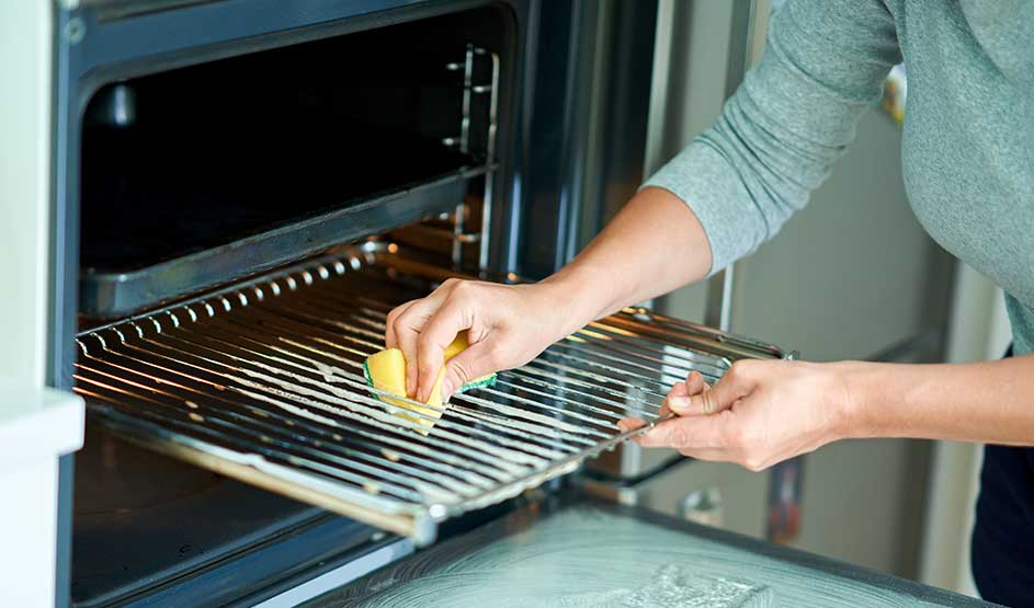 Iemand maakt het ovenrooster schoon met een spons terwijl de ovendeur openstaat in een heldere keuken.