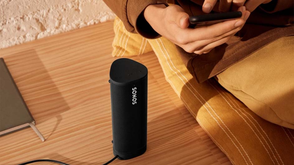 Een zwarte Sonos speaker staat op een houten vloer, naast een persoon met een telefoon en een notitieboek.