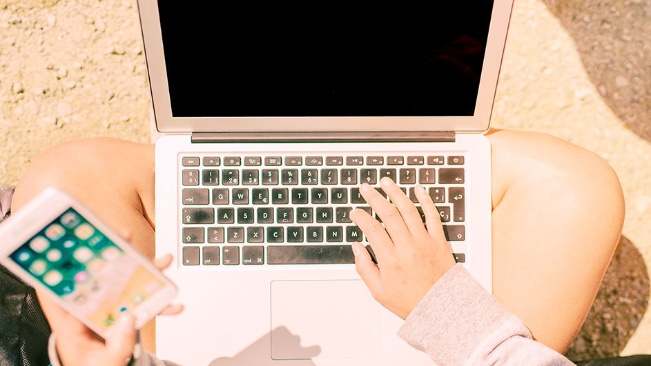 Person types on a laptop with a black screen, holding a smartphone with a colorful interface. The scene is outdoors on a light brown ground.