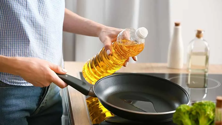 Een persoon giet olie uit een plastic fles in een zwarte koekenpan op een fornuis, met groenten op de voorgrond.