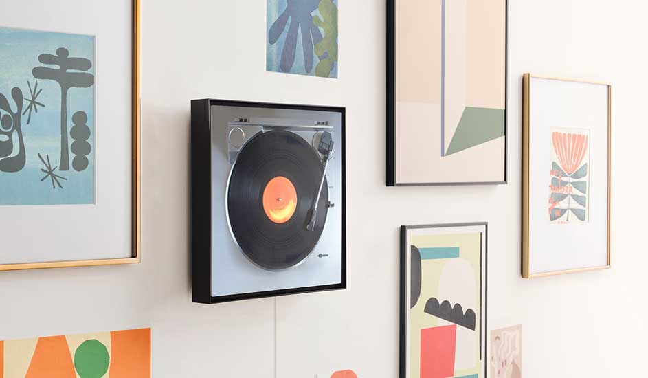 Een platenspeler hangt aan de muur tussen kleurrijke ingelijste kunstwerken, waardoor het lijkt op een kunstobject in een moderne woonkamer.