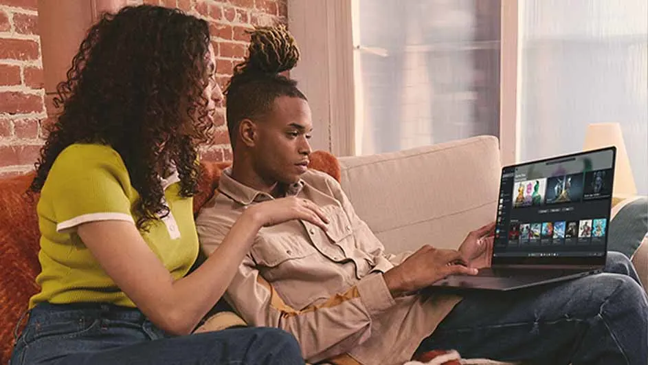 Twee mensen zitten op een bank naar een laptop te kijken. Achtergrond is een bakstenen muur.