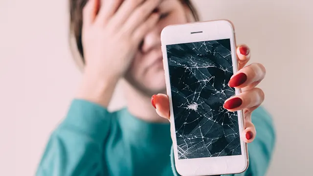 Een persoon met een blauw shirt houdt een witte telefoon vast met een gebarsten scherm. De achtergrond is lichtroze.