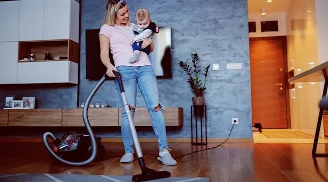 A mother vacuums the living room while holding her baby, both smiling above the rug on a cosy wooden floor.