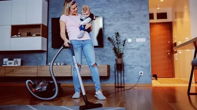 A mother vacuums the living room while holding her baby, both smiling above the rug on a cosy wooden floor.