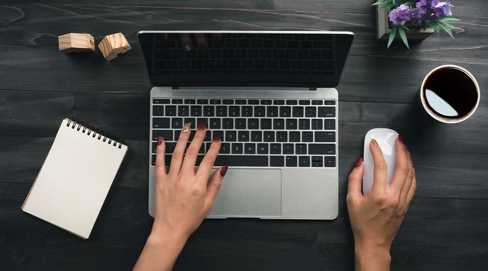 Overhead view of a laptop in gebruik, met hands on keyboard and mouse, flanked by a notebook, coffee mug and small plant.