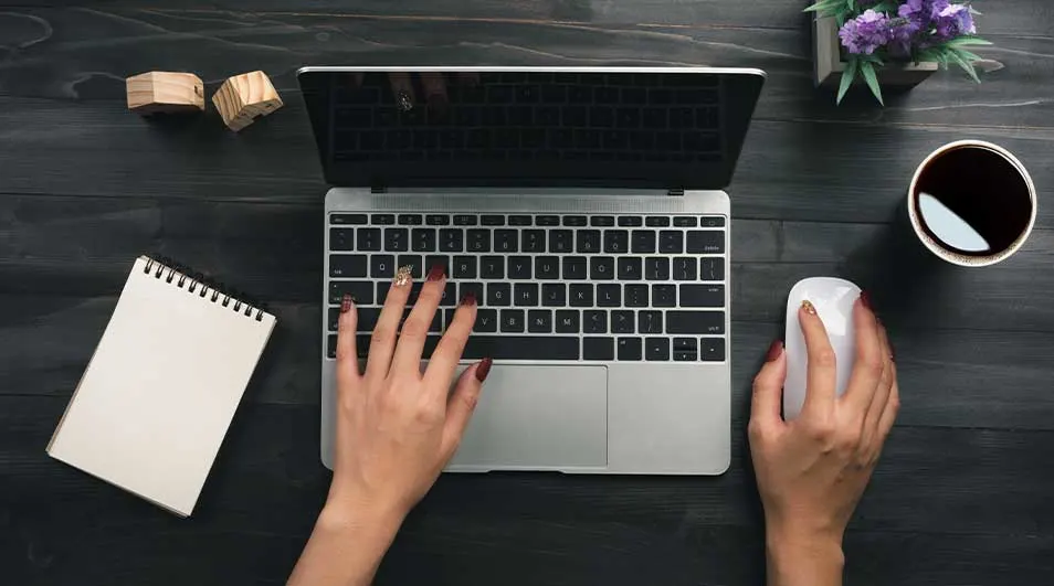 Overhead view of a laptop in gebruik, met hands on keyboard and mouse, flanked by a notebook, coffee mug and small plant.