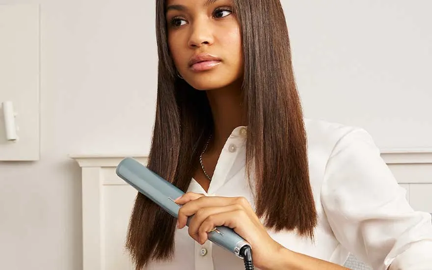 A young woman with long, straight hair holds a hair straightener in a bright room, her style looking smooth and polished.