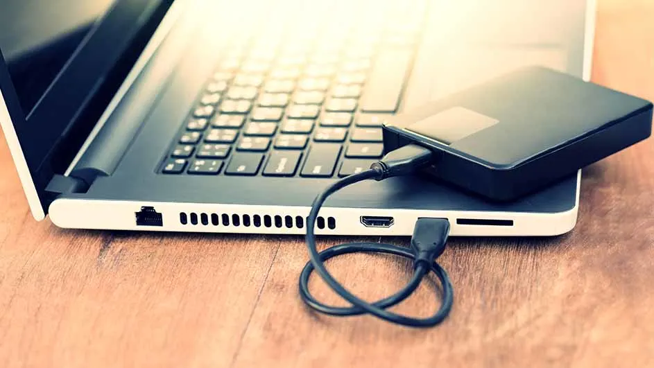 Een laptop met een zwarte externe harde schijf erop, verbonden met een zwarte kabel op een houten tafel.