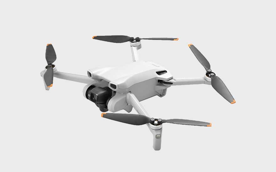 Op de afbeelding staat een compacte, lichtgrijze drone met opvallende oranje accenten op de propellerpunten. 