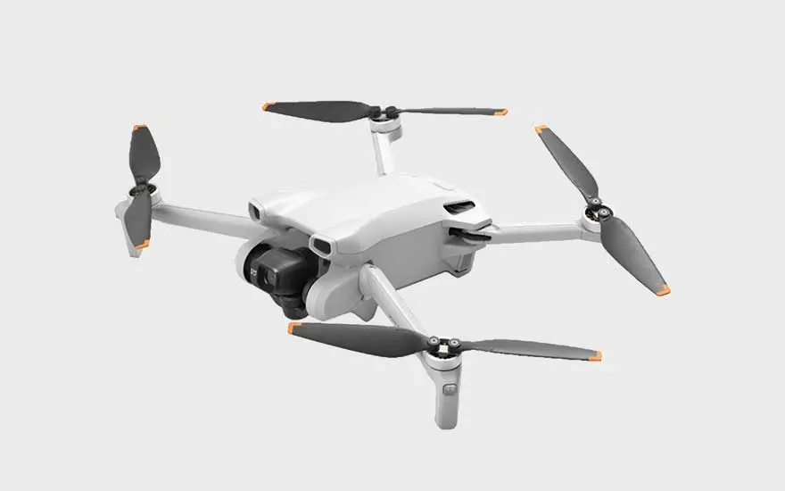 Op de afbeelding staat een compacte, lichtgrijze drone met opvallende oranje accenten op de propellerpunten.