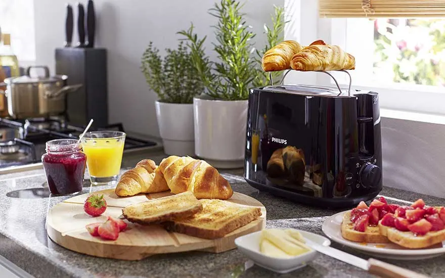 Een zwarte Philips broodrooster met twee croissants erboven, naast een rijk ontbijtbord met toast, croissants, aardbeien, confiture en een glas jus d’orange.
