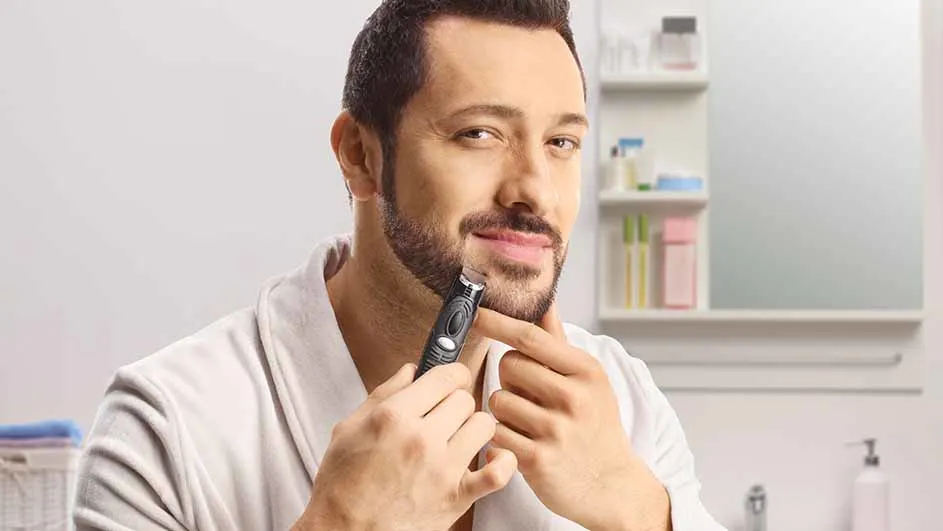 Man in badjas trimt baard met een zwart scheerapparaat in een badkamer met witte muren en een spiegelkast.