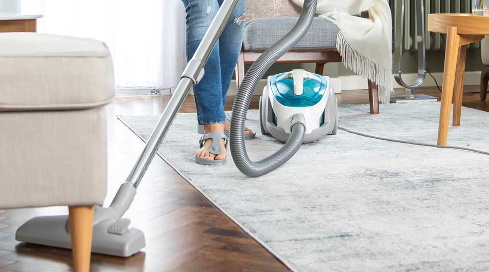 A person hoovers a pale rug in a bright living room, with wooden furniture, soft textiles and a compact cylinder vacuum cleaner.