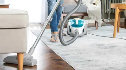 A person hoovers a pale rug in a bright living room, with wooden furniture, soft textiles and a compact cylinder vacuum cleaner.