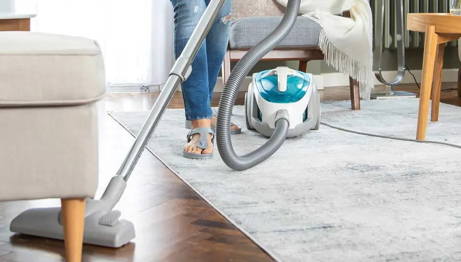 Een persoon stofzuigt een licht vloerkleed in een gezellige woonkamer, met een modern tapijt, houten meubels en een compacte stofzuiger.