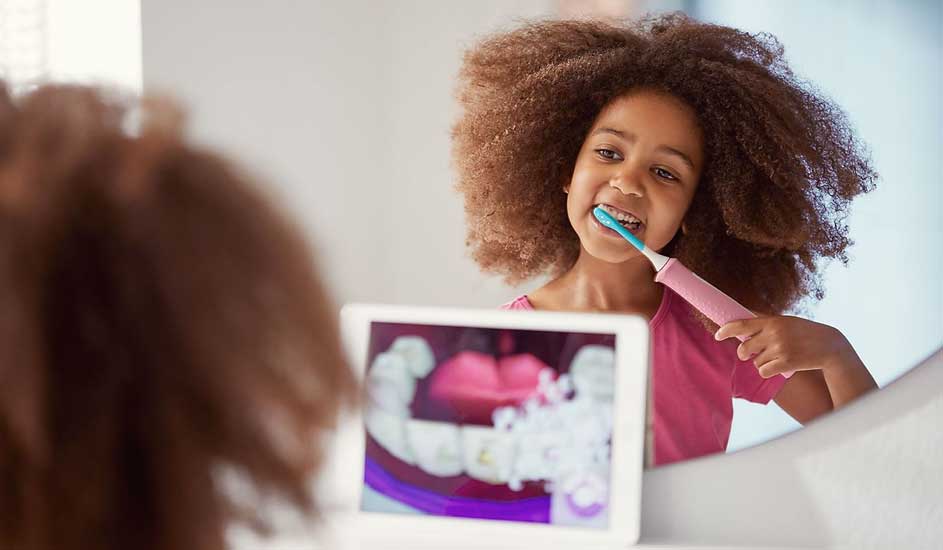 Een kind met een grote bos krullen borstelt het haar met een roze borstel, terwijl op een tablet een animatie van tandenpoetsen te zien is.