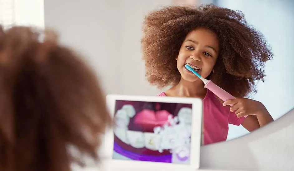 Een kind met een grote bos krullen borstelt het haar met een roze borstel, terwijl op een tablet een animatie van tandenpoetsen te zien is.