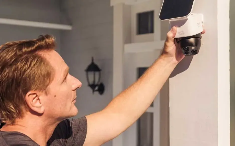 Een man monteert een witte beveiligingscamera met zwart paneel en zonnepaneel van het merk Ezviz aan een buitenmuur van een huis.