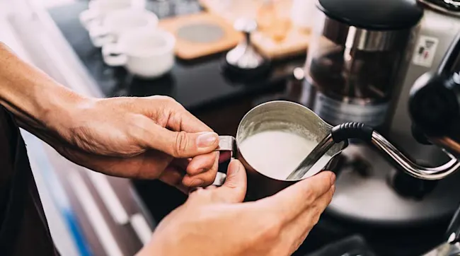 Hands hold a metal pitcher of white milk by an espresso machine. Cups and a wooden board are in the background.
