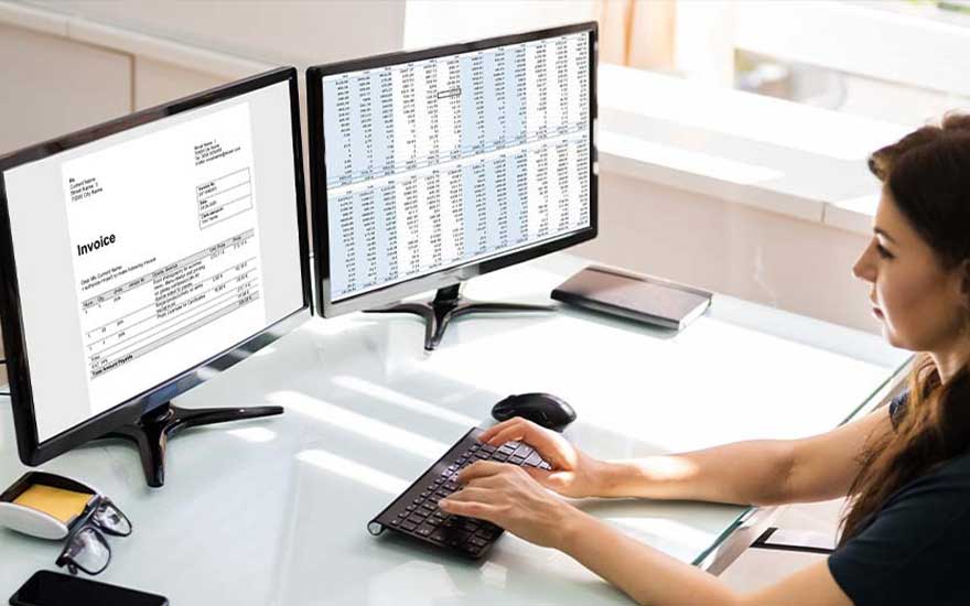 A woman uses dual monitors showing an invoice and spreadsheet, typing on a compact keyboard in a bright, minimalist home office.