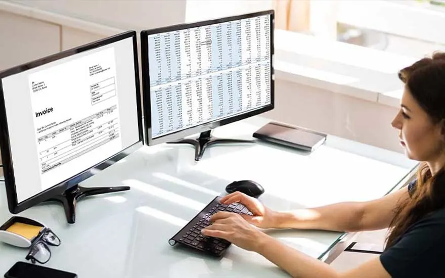 A woman uses dual monitors showing an invoice and spreadsheet, typing on a compact keyboard in a bright, minimalist home office.