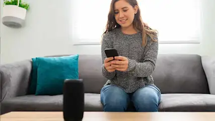 Een vrouw zit op een grijze bank met een blauw kussen en kijkt naar een telefoon. Een zwarte speaker staat op een houten tafel.