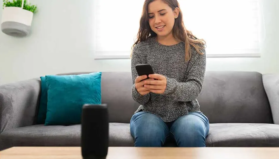 Een vrouw zit op een grijze bank met een blauw kussen en kijkt naar een telefoon. Een zwarte speaker staat op een houten tafel.