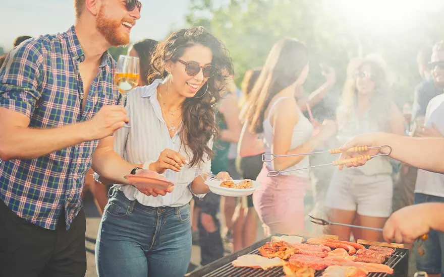 Friends laugh around a sunny barbecue, drinks in hand, while fresh meat sizzles on the hot grill in a lively setting.