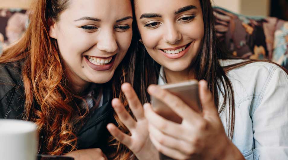 Twee jonge vrouwen lachen samen terwijl ze op een smartphone kijken; het scherm weerspiegelt hun vrolijke gezichten. Geen merk zichtbaar.